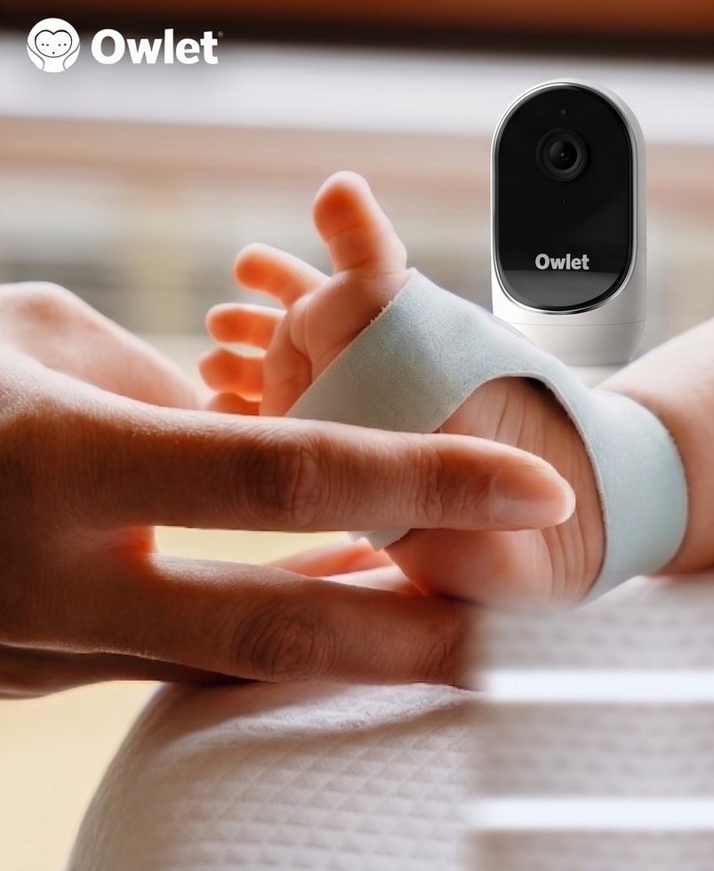Mâna unui părinte ținând piciorul bebelușului cu senzorul Owlet Dream Sock, alături de camera Owlet